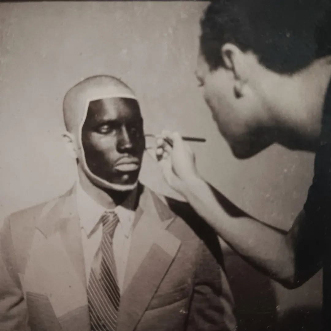 Black-and-white photograph of Andrei Scholz seated in a suit and tie with his head covered, makeup being applied to his face by a standing artist