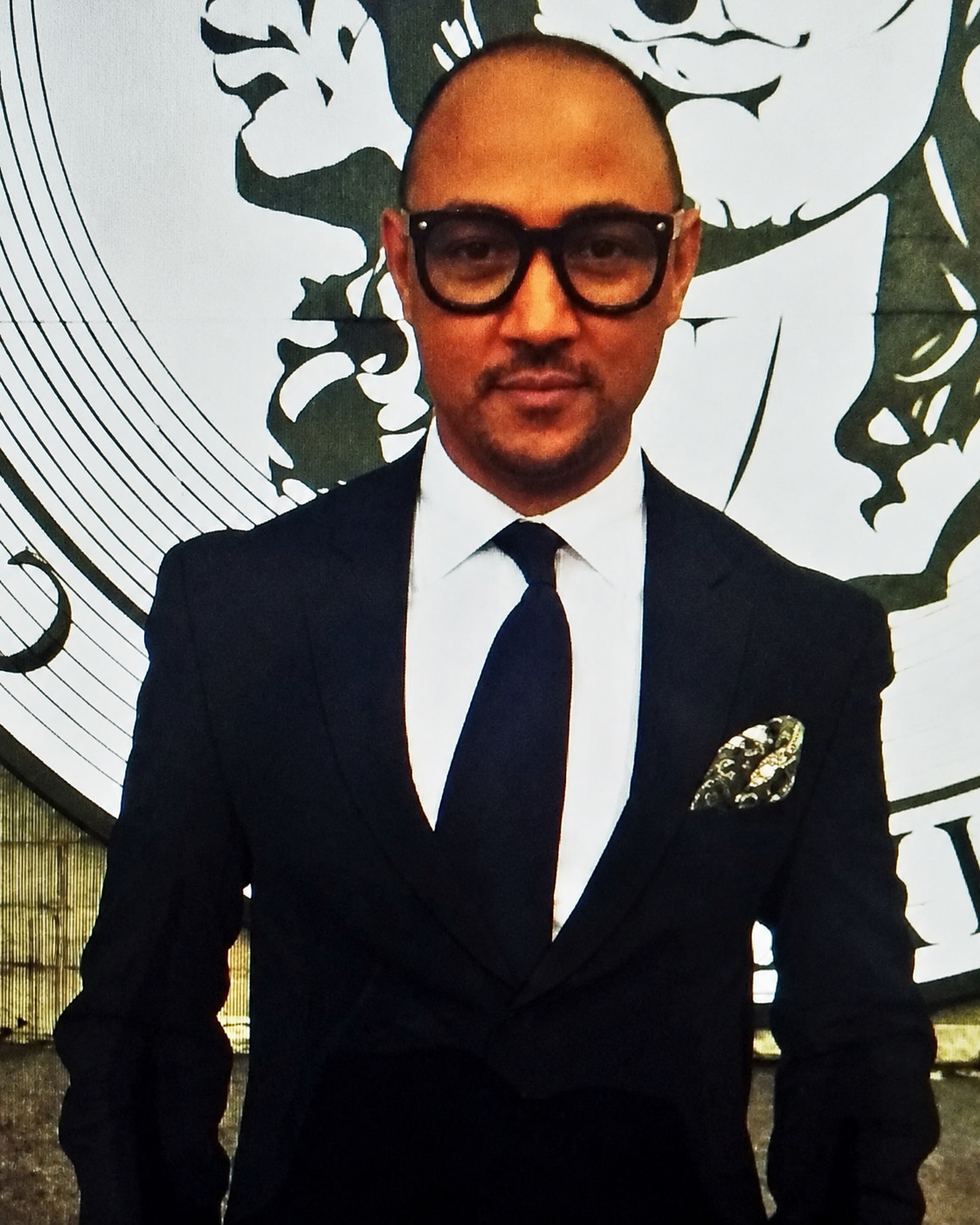 Andrei Scholz, makeup artist and founder of A.B.B.A., in a dark suit, tie and glasses against a graphic studio backdrop